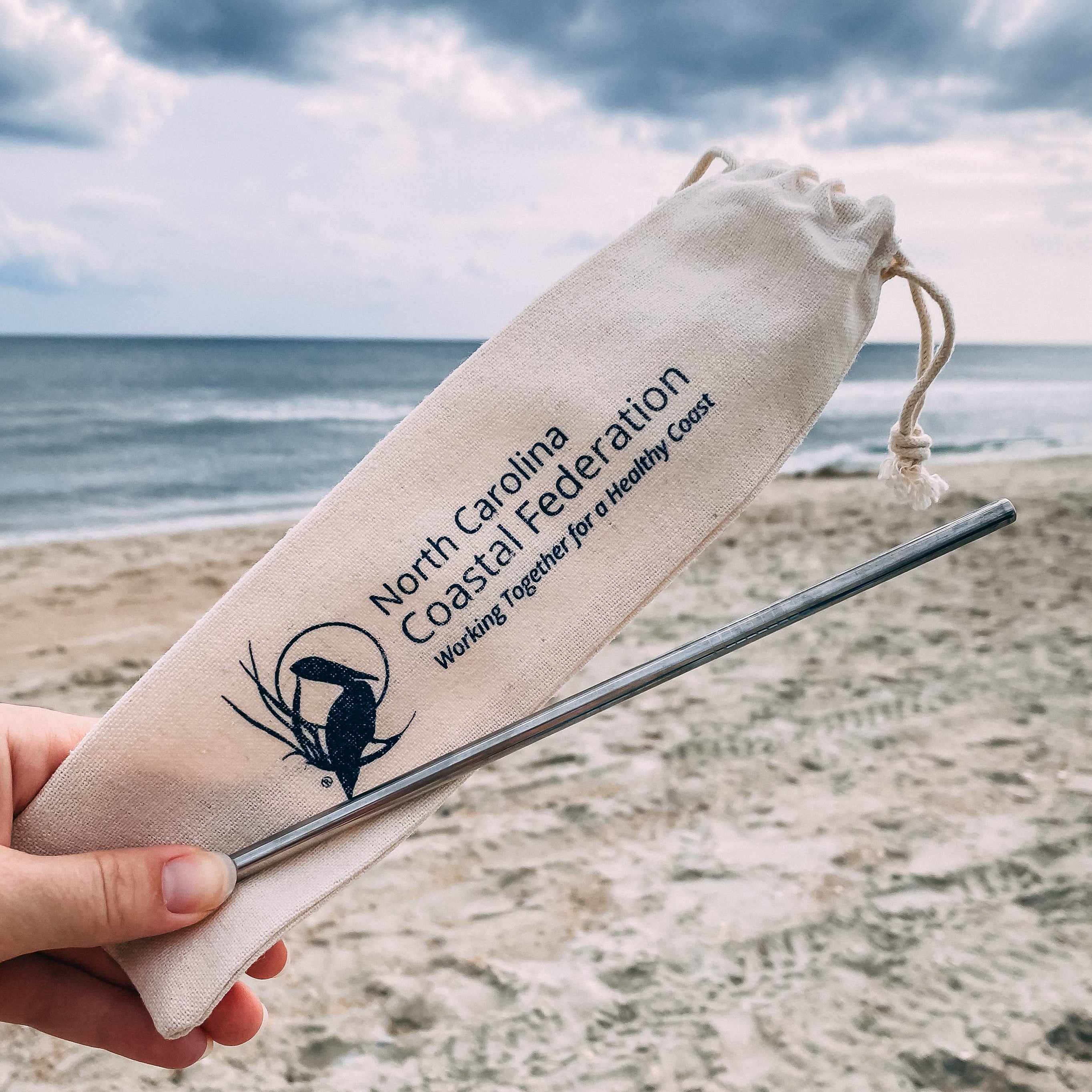 Straw and drawstring bag with North Carolina Coastal Federation logo on a beach.